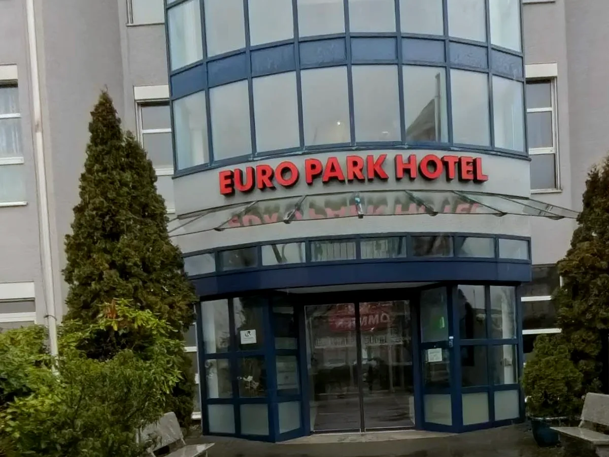 Haupteingang des EURO PARK HOTEL Hennef mit Schriftzug über dem Vordach, verglastem Treppenturm, Bänken und Grünpflanzen an einem trüben Tag
