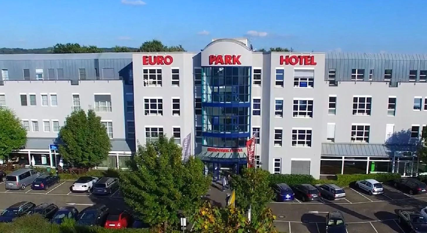Aerial view of the EURO PARK HOTEL Hennef front facade with red lettering, glazed stair tower and car park on a sunny day
