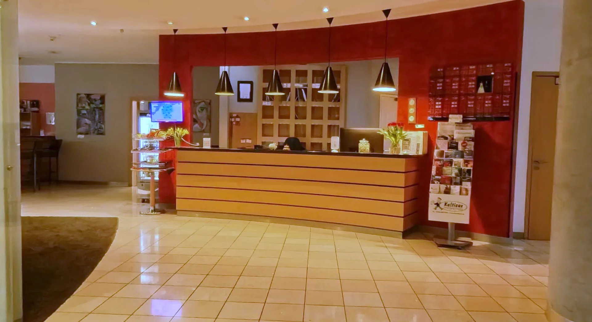 Rezeption im EURO PARK HOTEL Hennef mit geschwungener Holztheke vor roter Wand, schwarzen Hängeleuchten, Blumen und hellem Fliesenboden