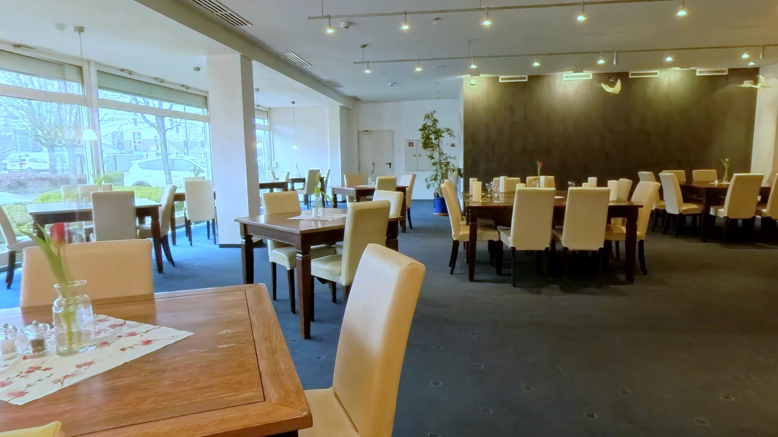 Restaurant im EURO PARK HOTEL Hennef mit Holztischen, cremefarbenen Stühlen, blauem Teppich, Tulpen in Glasvasen und großer Fensterfront