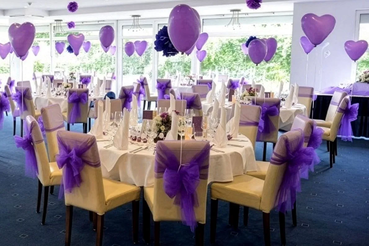 Hochzeitsdekoration im EURO PARK HOTEL Hennef: runde Tische mit weißen Decken, Stühle mit lila Schleifen und lila Herzballons vor großen Fenstern