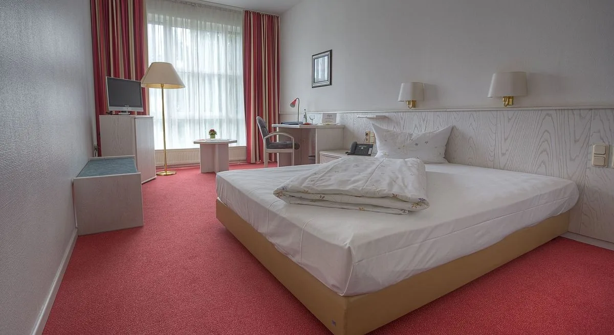 Businesszimmer im EURO PARK HOTEL Hennef mit Bett vor heller Holzvertäfelung, Schreibtisch am Fenster, Stehlampe, Fernseher und rotem Teppichboden