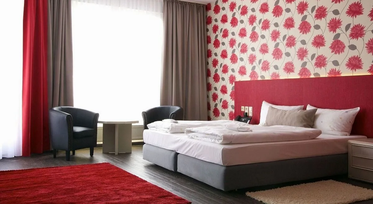 Komfortzimmer im EURO PARK HOTEL Hennef: Doppelbett mit rotem Kopfteil vor Blumentapete, zwei dunkle Sessel am Fenster und roter Teppich