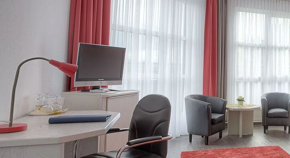 Schreibtisch mit roter Schreibtischlampe und Freischwinger im Komfortzimmer des EURO PARK HOTEL Hennef, daneben Fernseher, zwei Sessel und helle Fenster