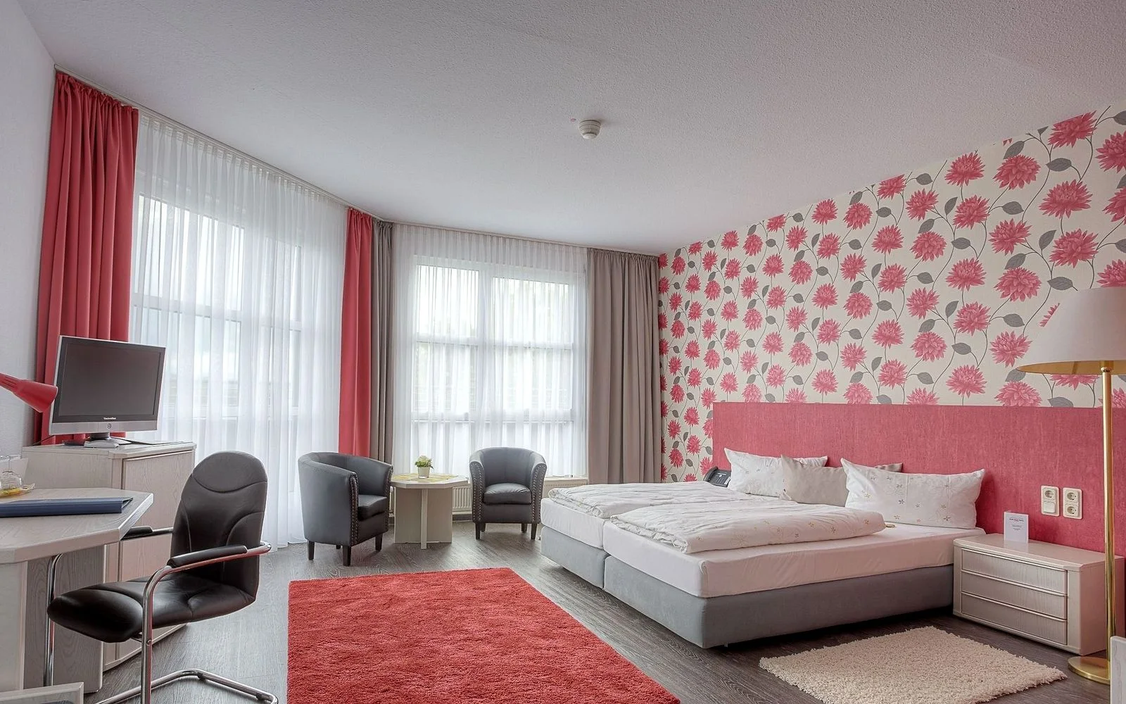 Komfortzimmer im EURO PARK HOTEL Hennef mit Schreibtisch, zwei Sesseln am Fenster, Doppelbett vor Tapete mit roten Blüten und rotem Teppich