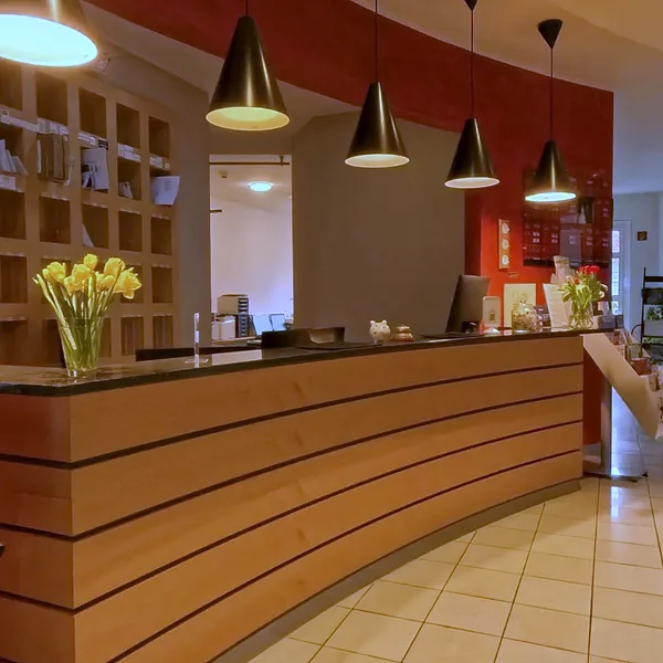 Rezeption im EURO PARK HOTEL Hennef: geschwungene Holztheke vor roter Wand, schwarze Hängeleuchten und gelbe Blumen auf dem Tresen