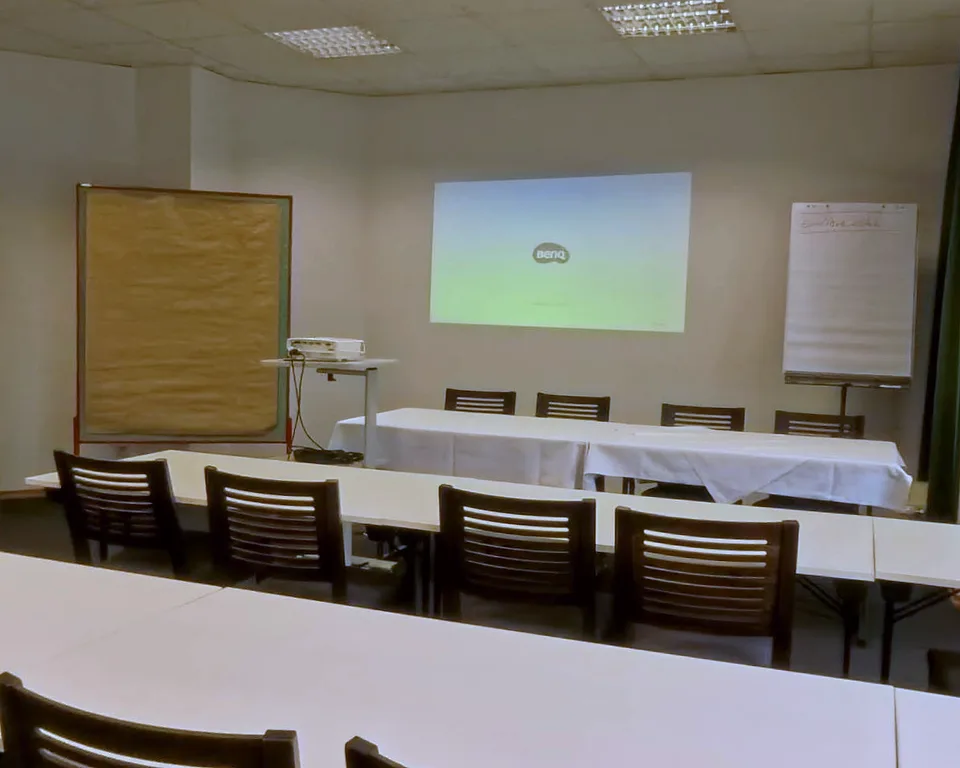 Projector on a rolling stand with an image on the wall, a pinboard and a flipchart in the Hennef room at EURO PARK HOTEL Hennef, white tables in front