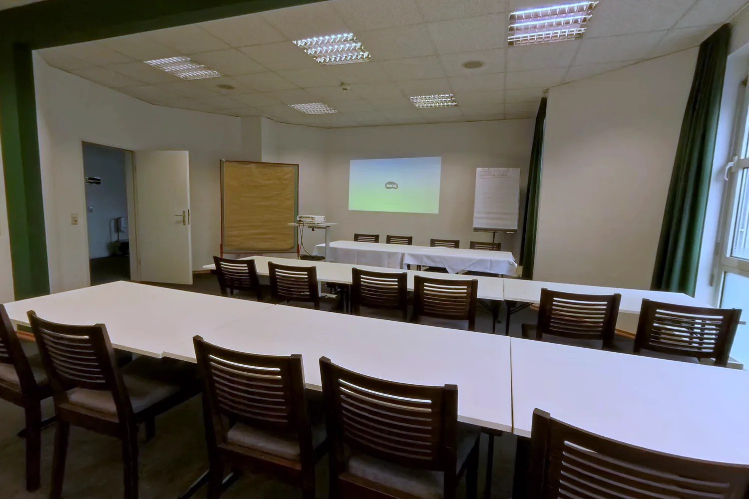 Hennef room at EURO PARK HOTEL Hennef: white table rows with dark wooden chairs, a projected image on the wall, projector, pinboard and flipchart