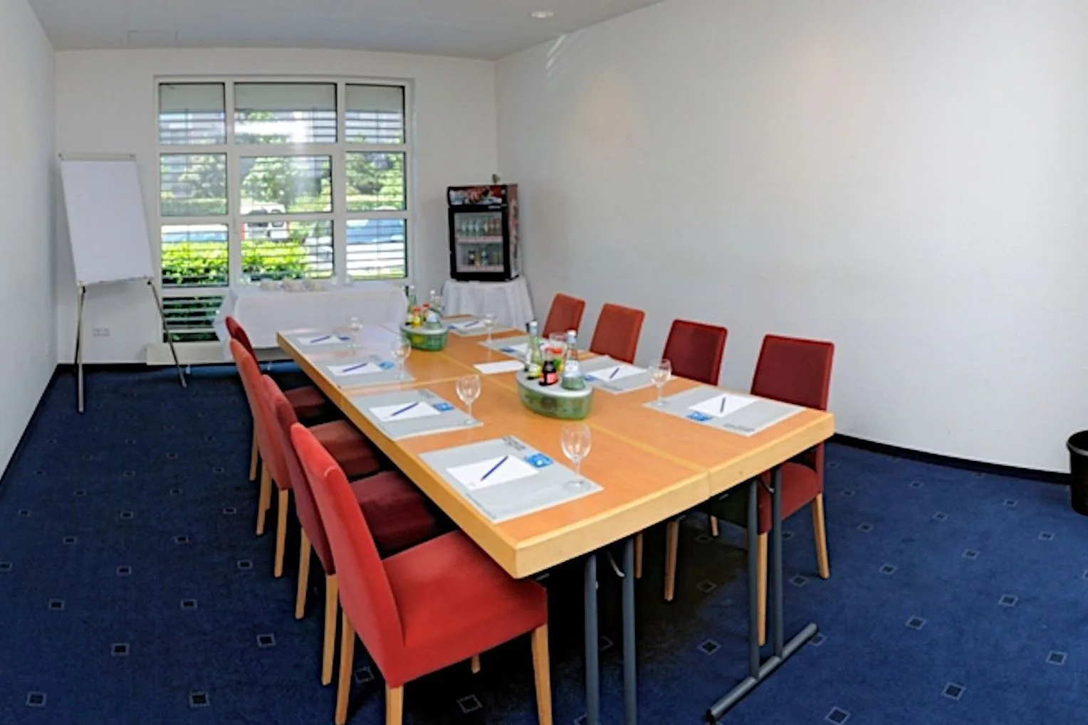 Sankt Augustin meeting room at EURO PARK HOTEL Hennef: wooden table with red upholstered chairs, notepads, a flipchart, a mini fridge and a window