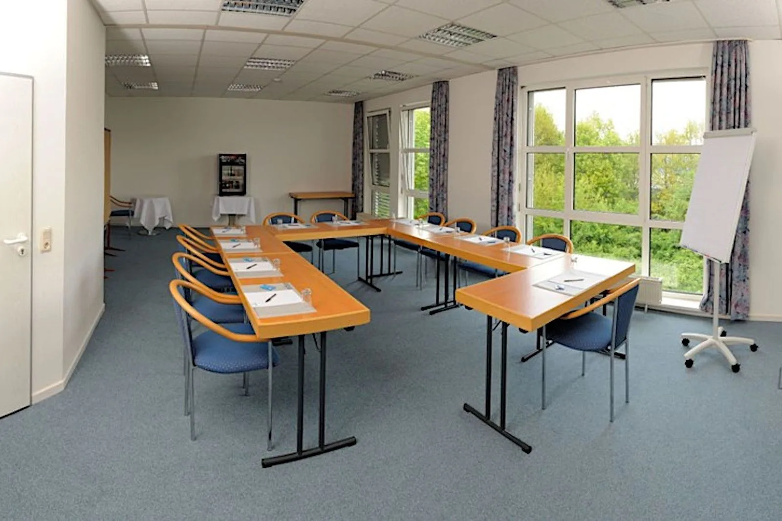 Troisdorf meeting room at EURO PARK HOTEL Hennef: U-shaped light wood tables with blue chairs, a flipchart, patterned curtains and windows onto trees