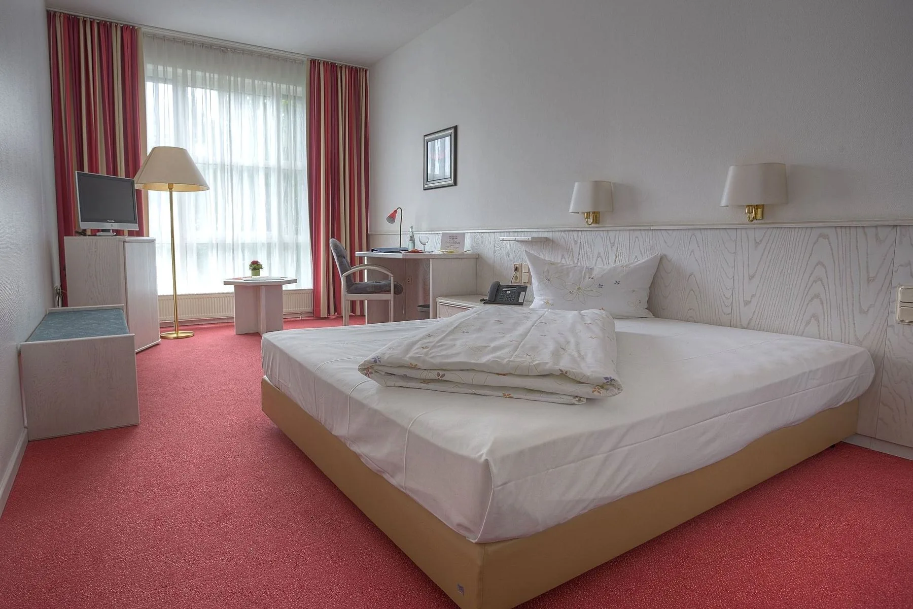Business room at EURO PARK HOTEL Hennef with a wide bed, wall lamps, a desk and chair, a floor lamp by the window and red fitted carpet