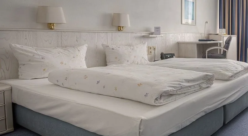 Doppelbett mit zwei Kissen, gefalteten Bettdecken und Wandleuchten in einem Gästezimmer des EURO PARK HOTEL Hennef, dahinter der Schreibtisch