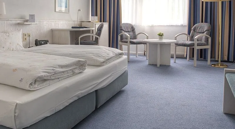 Blick über Bett und blauen Teppichboden zur Sitzecke mit zwei Sesseln, Tisch und Stehlampe in einem Zimmer des EURO PARK HOTEL Hennef