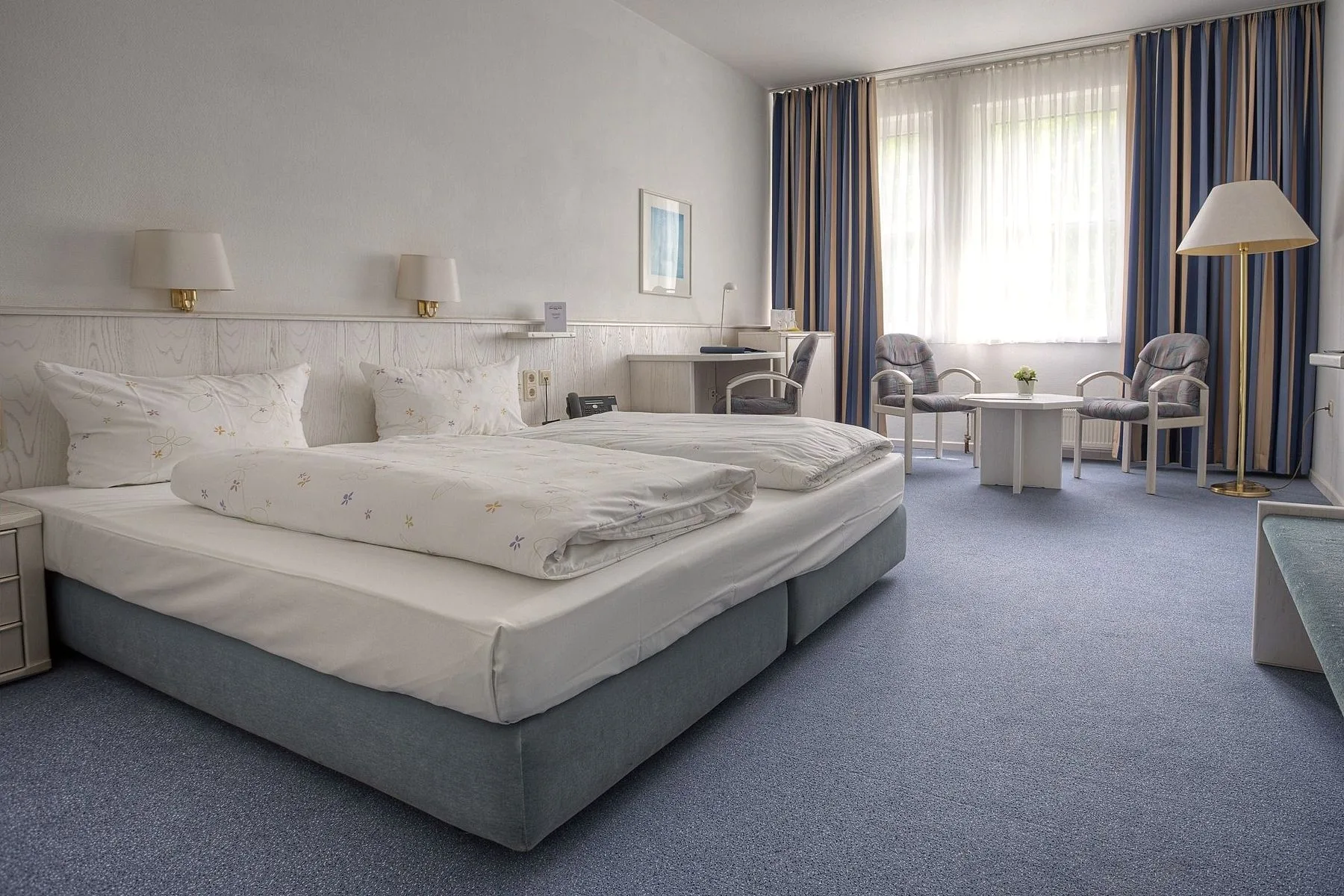 Standardzimmer im EURO PARK HOTEL Hennef mit Doppelbett, hellem Holzpaneel, Schreibtisch, zwei Sesseln am Tisch und blauem Teppichboden